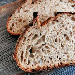 Zwei Scheiben rustikales Sauerteigbrot mit großer Porung liegen auf einem Holzbrett. Warum ist Sauerteig gesund? Weil die Fermentation Nährstoffe besser verfügbar macht.