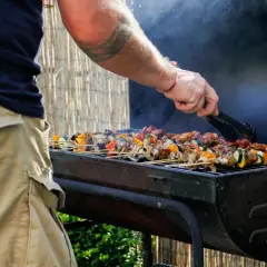 Un homme utilise des pinces pour retourner des brochettes colorées sur un barbecue fumant. C'est le moment idéal pour degraisser le barbecue après ce festin !