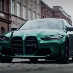 Zielony BMW M3 Competition z charakterystycznym grillem, zaparkowany na brukowanej ulicy przed ceglanymi budynkami.