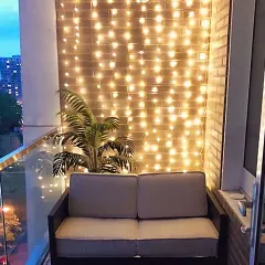 Przytulny balkon w bloku z sofą i palmą, ozdobiony ciepłym oświetleniem.