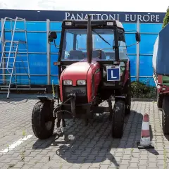 Czerwony traktor z literą "L" (kategoria t prawo jazdy) stoi obok przyczepy. W tle niebieska ściana i drabina.