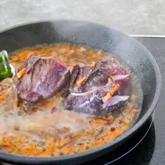 Deux morceaux de joue de bœuf mijotent dans une poêle avec des carottes et des oignons. Un liquide est versé d'une bouteille verte.