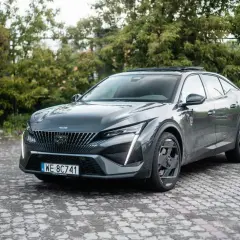 Elegancki, ciemnoszary Peugeot 408 GT z charakterystycznymi światłami do jazdy dziennej stoi na brukowanym podjeździe, otoczony zielenią.