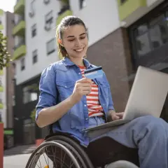 Kobieta na wózku inwalidzkim z laptopem i kartą kredytową cieszy się zakupami online, pokazując, że znaczny stopień niepełnosprawności nie ogranicza jej możliwości.