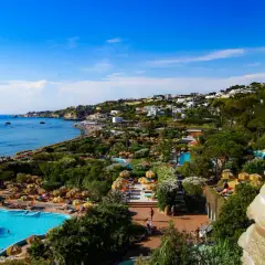 Vista panoramica dell'isola d'Ischia con mare azzurro, piscine, vegetazione lussureggiante e case arroccate sulla collina.