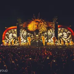 Nocny hardstyle festival z ogromną sceną i tłumem ludzi. W tle świecąca, złota głowa w koronie.