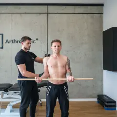 Fizjoterapeuta pomaga pacjentowi w ćwiczeniach rehabilitacja barku po operacji. Mężczyzna trzyma kij, wykonując ruchy ramionami.