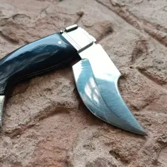 Navaja capaora de mango negro y hoja curva, lista para la acción sobre una superficie rocosa.