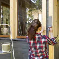 Kvinna målar i solsken en träpanel på ett hus. En stege och en färgburk står bredvid.