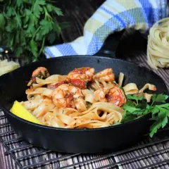 Spaghetti mit Garnelen mediterran in einer Gusseisenpfanne, garniert mit Zitrone und Petersilie. Daneben rohe Pasta, Knoblauch und Chili.