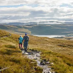 Two hikers trek along the Cape Wrath Trail, a rugged path winding through a vast, misty Scottish landscape with a serene loch in the distance.