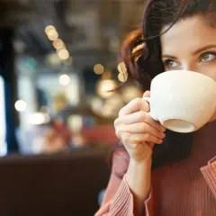 En kvinna med mörkt hår dricker kaffe ur en vit kopp. Hon har en stickad tröja och sitter på ett café. Kanske lite kaffe på tom mage för att vakna till.