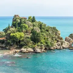 En grönskande ö med klippor och byggnader, omgiven av turkost vatten. En smal sandremsa leder till ön, där människor solar och badar. En idyllisk vy av **white lotus sicily**.