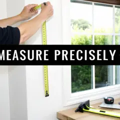 A person measures a window frame, demonstrating how to measure for pinch pleat curtains. Tools are laid out on a wooden surface.