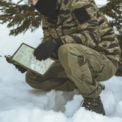 Osoba w zimowym kamuflażu i spodniach Helikon M65 analizuje mapę w śnieżnym otoczeniu.