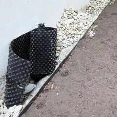 Schwarze Noppenbahn schützt die Hauswand vor Feuchtigkeit, daneben Kies und Erde.