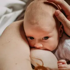 Spokojny noworodek przy piersi, z otwartymi oczami, to znak, po czym poznać, że noworodek jest najedzony. Dłoń czule głaszcze jego główkę.