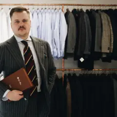 Elegancki mężczyzna w garniturze w prążki trzyma teczkę. Za nim wiszą marynarki i koszule.
