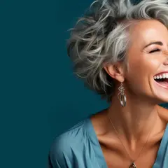 Frau mit vollem Lachen und auffälligen blau grauen Haaren, die im Wind wehen. Sie trägt einen blauen Seidenstoff und auffälligen Schmuck.