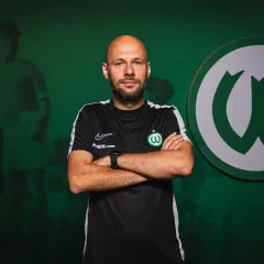 Łysy mężczyzna z brodą, w czarnej koszulce z zielonym herbem, stoi z rękami skrzyżowanymi na piersi. To trener Warty Poznań.