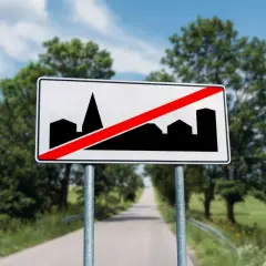 Znak d43: koniec obszaru zabudowany. Droga prowadzi przez zielone krajobrazy, a znak informuje o końcu miasta.