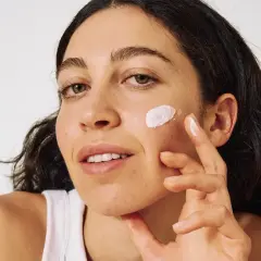 Frau trägt sanft Sonnencreme auf ihre Wange auf, um ihre Haut vor Akne zu schützen.