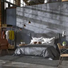 Przytulny soft loft z łóżkiem, fotelem i biurkiem. Duże okna wpuszczają światło, podkreślając surowy charakter wnętrza.