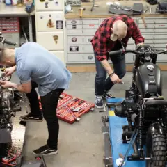 Due uomini lavorano su due moto in un garage. Uno è chino su una moto bianca, l'altro su una nera. Un kit di attrezzi è aperto sul pavimento. Progetto schienalino moto fai da te.
