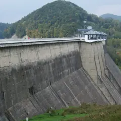 Ogromna zapora na sanie góruje nad zielonymi wzgórzami. Na jej szczycie widać ludzi spacerujących po moście.