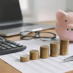 Kalkulator, laptop, okulary, skarbonka i rosnące stosy monet na dokumentach finansowych. Obrazek symbolizuje zarządzanie finansami i potencjalny koszt pracodawcy.