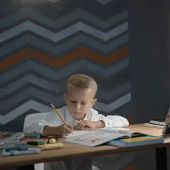 Chłopiec pisze przy biurku, które ma odpowiednie wymiary biurka dla dziecka. Na blacie leżą książki, tablet i lampka.
