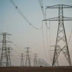 Linia wysokiego napięcia z wieżami energetycznymi, przypominającymi gigantyczne kogeneratory, ciągnie się przez pustynny krajobraz.
