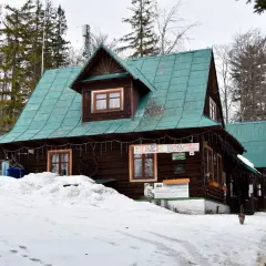 Drewniany domek z zielonym dachem, otoczony śniegiem i lasem. Na ścianie wisi baner "BĘDZIE DOBRZE", a przed wejściem stoi kot. Taki widok to prawdziwy przegibek!
