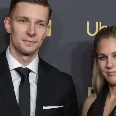 Aleksandra Mirosław i jej mąż, ubrani elegancko, pozują do zdjęcia na tle czarnej ściany z logo.