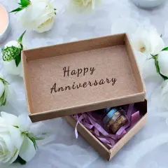 Otwarta szkatułka z napisem "Happy Anniversary" i małą buteleczką w środku, otoczona białymi różami i płonącą świecą. Idealny prezent na 21 rocznicę ślubu.