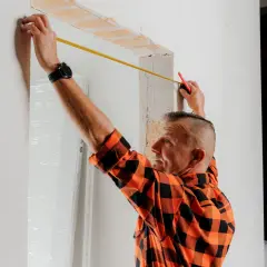 Budowlaniec w pomarańczowej koszuli mierzy taśmą otwór w ścianie.