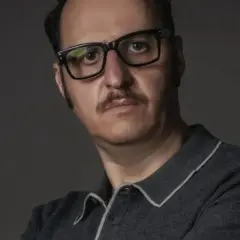 Ritratto di Fausto Russo Alesi con occhiali neri e baffi, indossa una polo grigia.