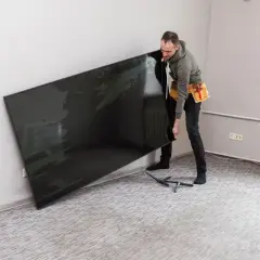 Mężczyzna z pasem narzędziowym przy ścianie trzyma duży, czarny telewizor. Zastanawia się, na jakiej wysokości telewizor będzie najlepiej wyglądał.