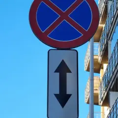Znak zakaz zatrzymywania z tabliczką wskazującą zakaz w obu kierunkach. Obok nowoczesny budynek z balkonami.
