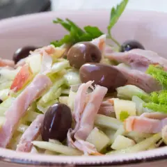 Frischer Fenchel Salat mit Apfelstücken, Schinkenstreifen und dunklen Oliven in einer rosa Schüssel.