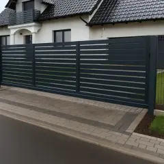Nowoczesna, ciemnoszara brama przesuwna, zapewniająca odpowiednią szerokość bramy wjazdowej dla tira, prowadzi do posesji z białym domem.