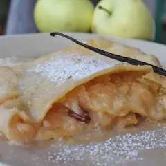 Ein Stück bayerischer Apfelstrudel, bestäubt mit Puderzucker und garniert mit einer Vanilleschote, daneben zwei Äpfel.