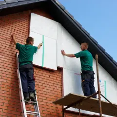 Deux ouvriers réalisent l'isolation par l'extérieur soi même d'une maison en briques, fixant des panneaux isolants blancs sur le pignon.