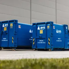 Dwa niebieskie kontenery BYS Recycling stoją przed budynkiem. Oferują tani transport odpadów.