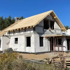 Budowa domu w lesie. Trwa montaż więźby dachowej, widać bloczki betonowe i przygotowania do instalacji. Wnętrza czekają na wykończenie.