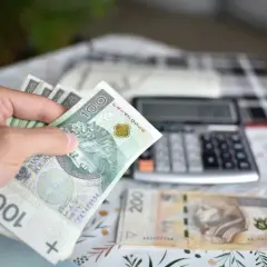 Ręka trzyma banknoty 100 zł, obok leży 200 zł i kalkulator. Obliczenia ekwiwalentu za urlop.