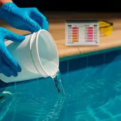Ręce w niebieskich rękawiczkach wlewają płyn do basenu. Obok tester wody, który pomoże określić, kiedy po chlorze szok można się kąpać.