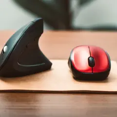 Dwa rodzaje mysz: ergonomiczna pionowa i klasyczna czerwona, leżące na drewnianym biurku.