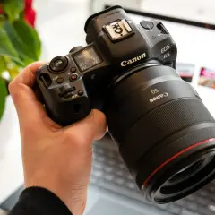 Dłoń trzyma aparat Canon EOS R5 z obiektywem 50mm. W tle widać ekran laptopa.