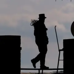 Silhouetten eines Bezirksschornsteinfegers mit Zylinder und Bürste auf dem Dach, der über ein Geländer geht.
